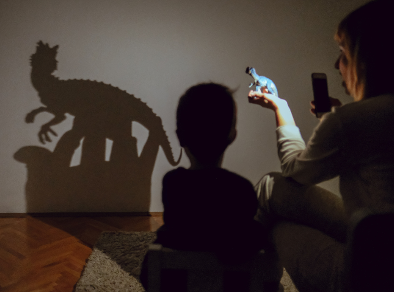 Dziecko i dorosły obserwują na ścianie cień figurki dinozaura oświetlonej latarką telefonu